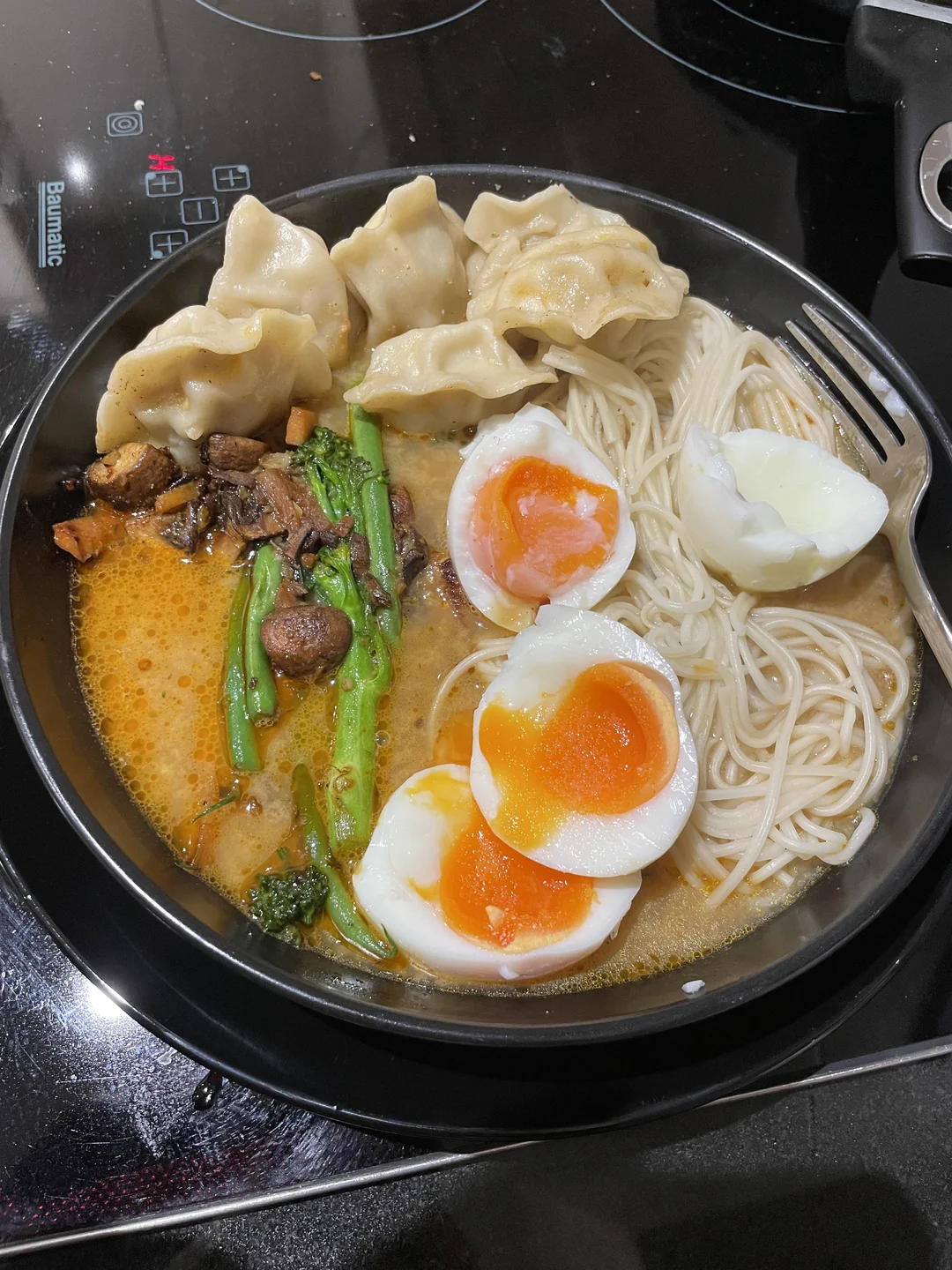 Bowl of ramen with soft-boiled eggs, noodles, greens, mushrooms, dumplings, and broth on a stovetop. A fork is resting on the side of the bowl.