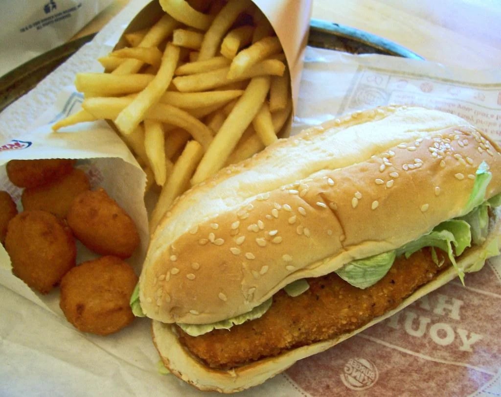A sesame seed sub sandwich with lettuce and breaded chicken, served with a side of French fries and several fried cheese bites on a wrapper.