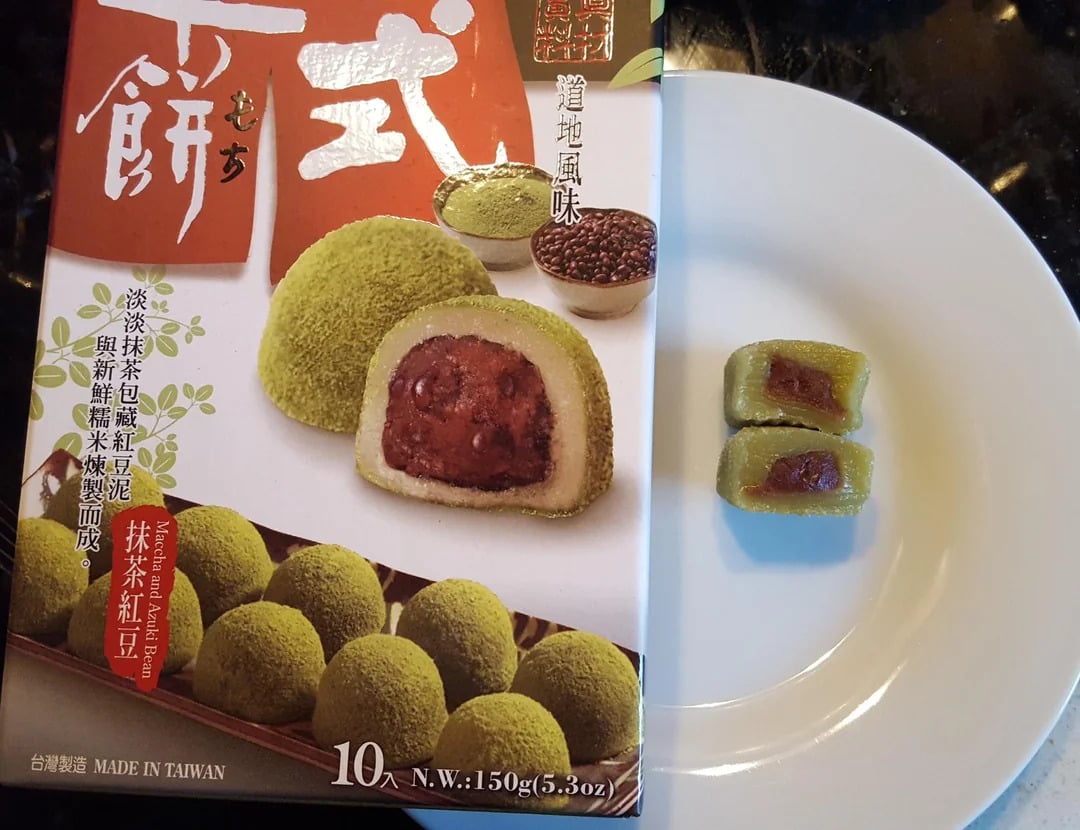 A box of matcha red bean mochi is shown next to a white plate with two cut pieces of the green mochi, revealing a red bean filling inside. The packaging displays images and text in Chinese and English.