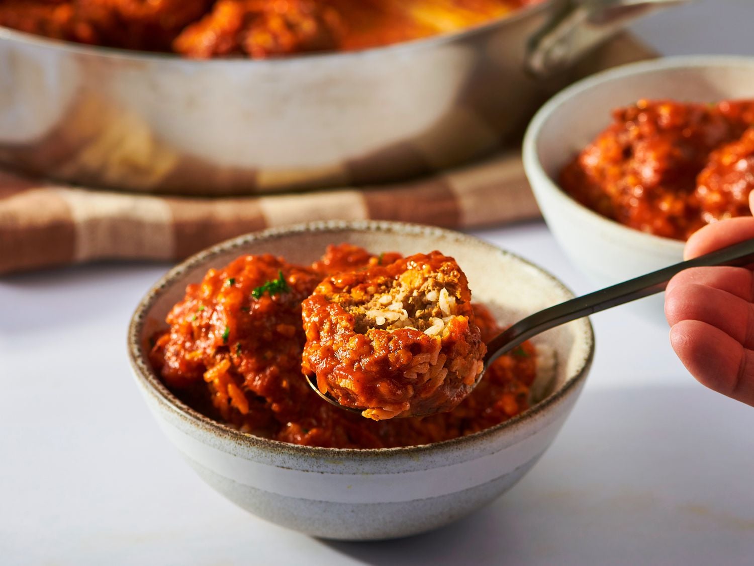 A hand holds a spoonful of saucy meatballs above a small bowl, with more meatballs and a pan in the background.