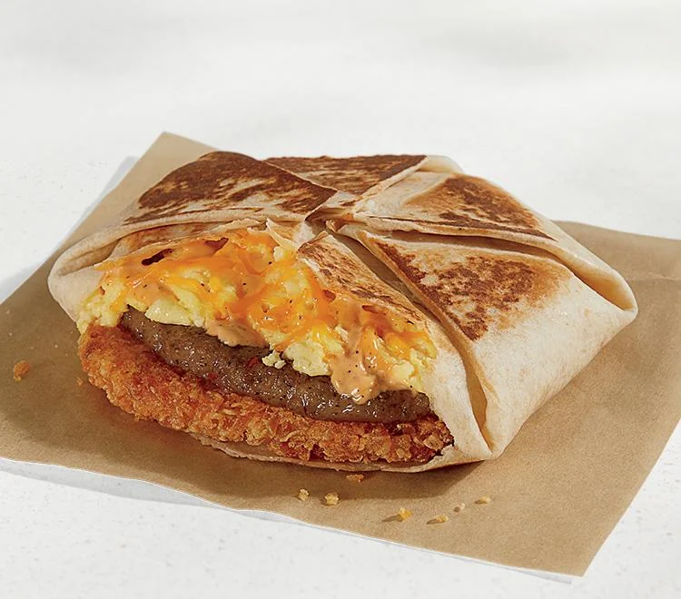 A folded toasted tortilla filled with scrambled eggs, sausage, cheese sauce, and a crispy hash brown, sitting on a piece of brown paper.