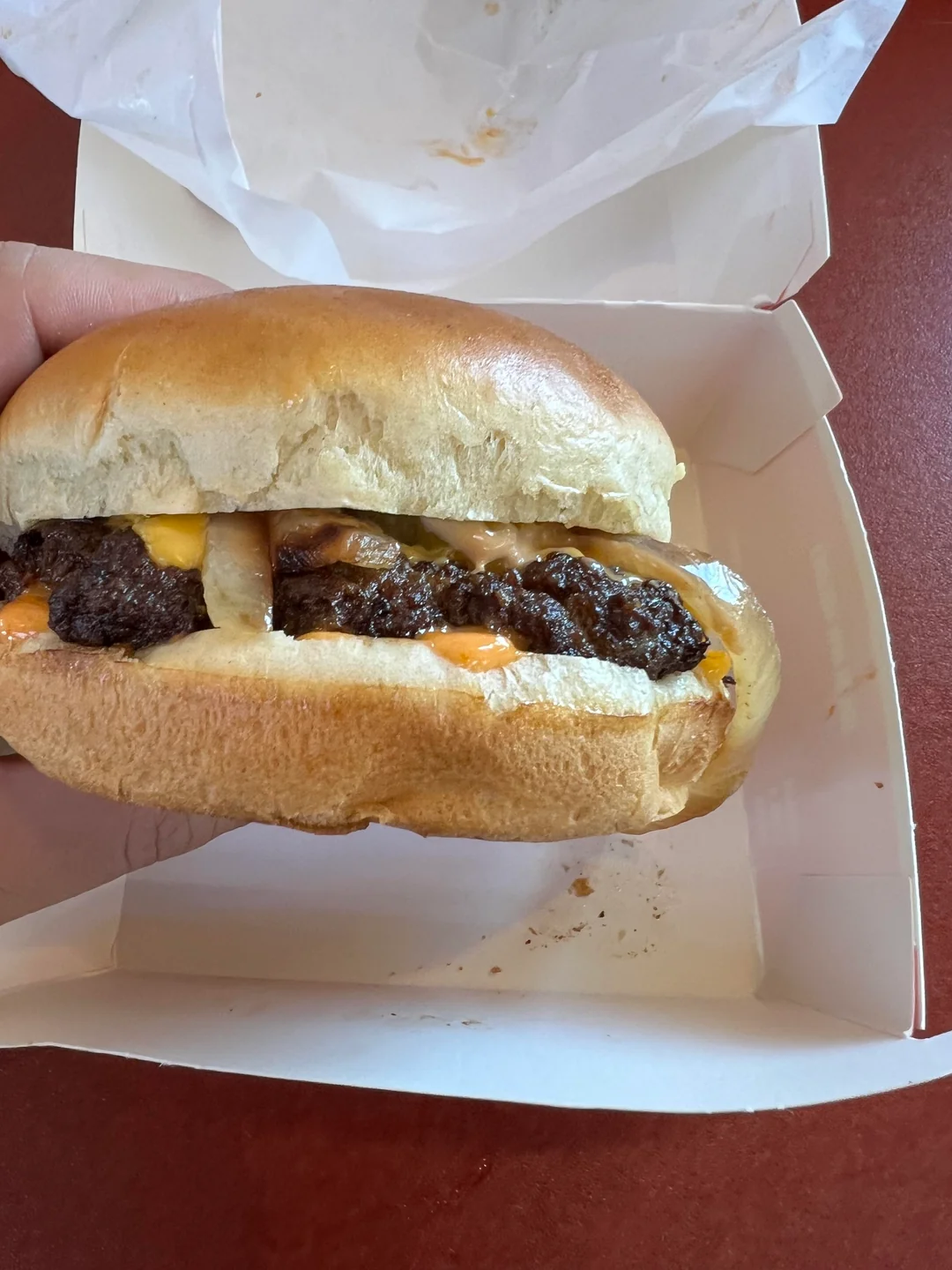 A hand holds a burger in a white takeout box. The burger has a beef patty, melted cheese, and sauce inside a soft bun, and the box rests on a reddish-brown table.
