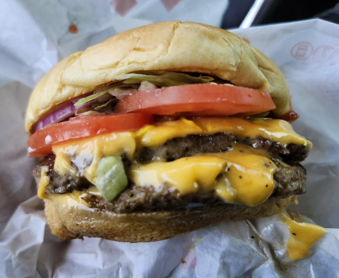 A cheeseburger with two beef patties, melted cheese, tomato slices, lettuce, pickles, onions, and a soft bun, wrapped in white paper.