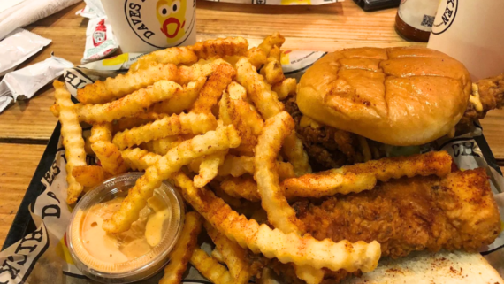Plate with crinkle-cut fries, a fried chicken sandwich on a bun, a fried chicken tender, and a small cup of dipping sauce&mdash;classic staples often featured at popular regional fast-food chains. A drink cup and condiment packets are visible in the background.