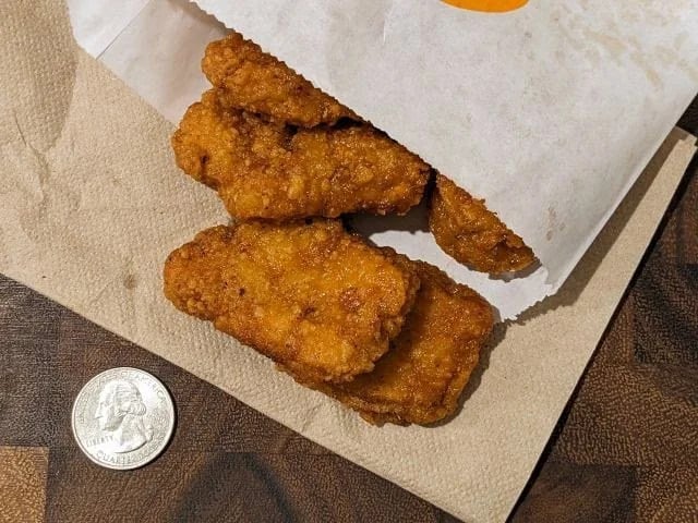 Four breaded chicken tenders spilling out of a white paper pouch onto a napkin, with a U.S. quarter placed nearby for size comparison. The items are on a wooden surface.