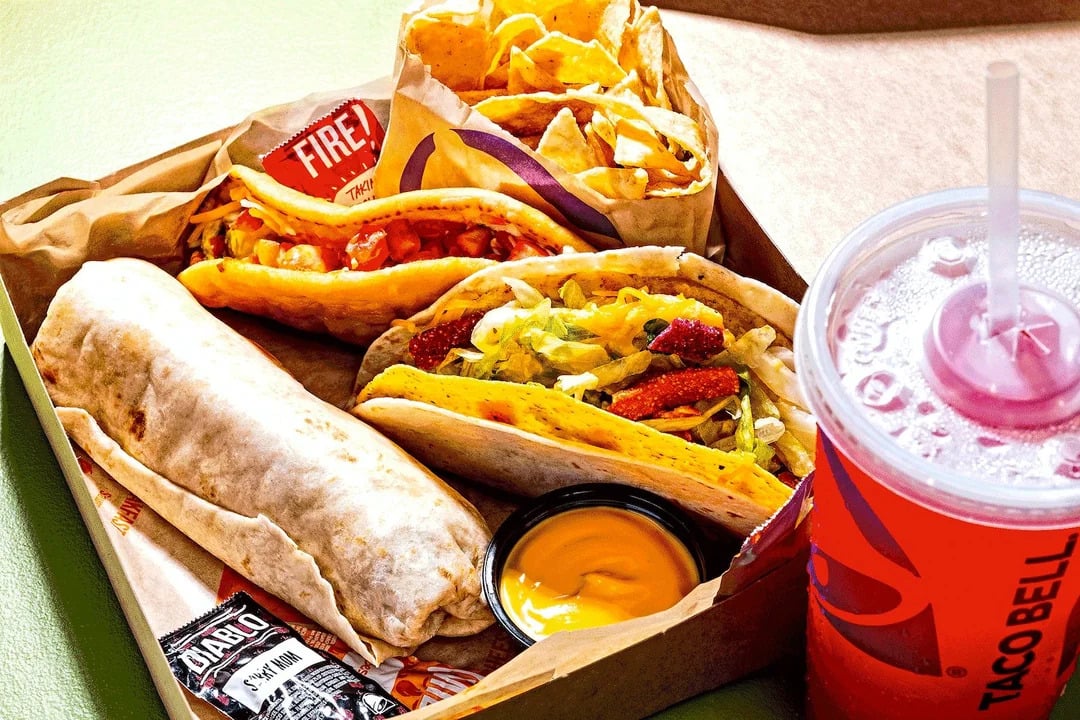 A Taco Bell meal with a burrito, two tacos, chips with cheese dip, hot sauce packets, and a large red drink with ice and a straw served in a takeout box.