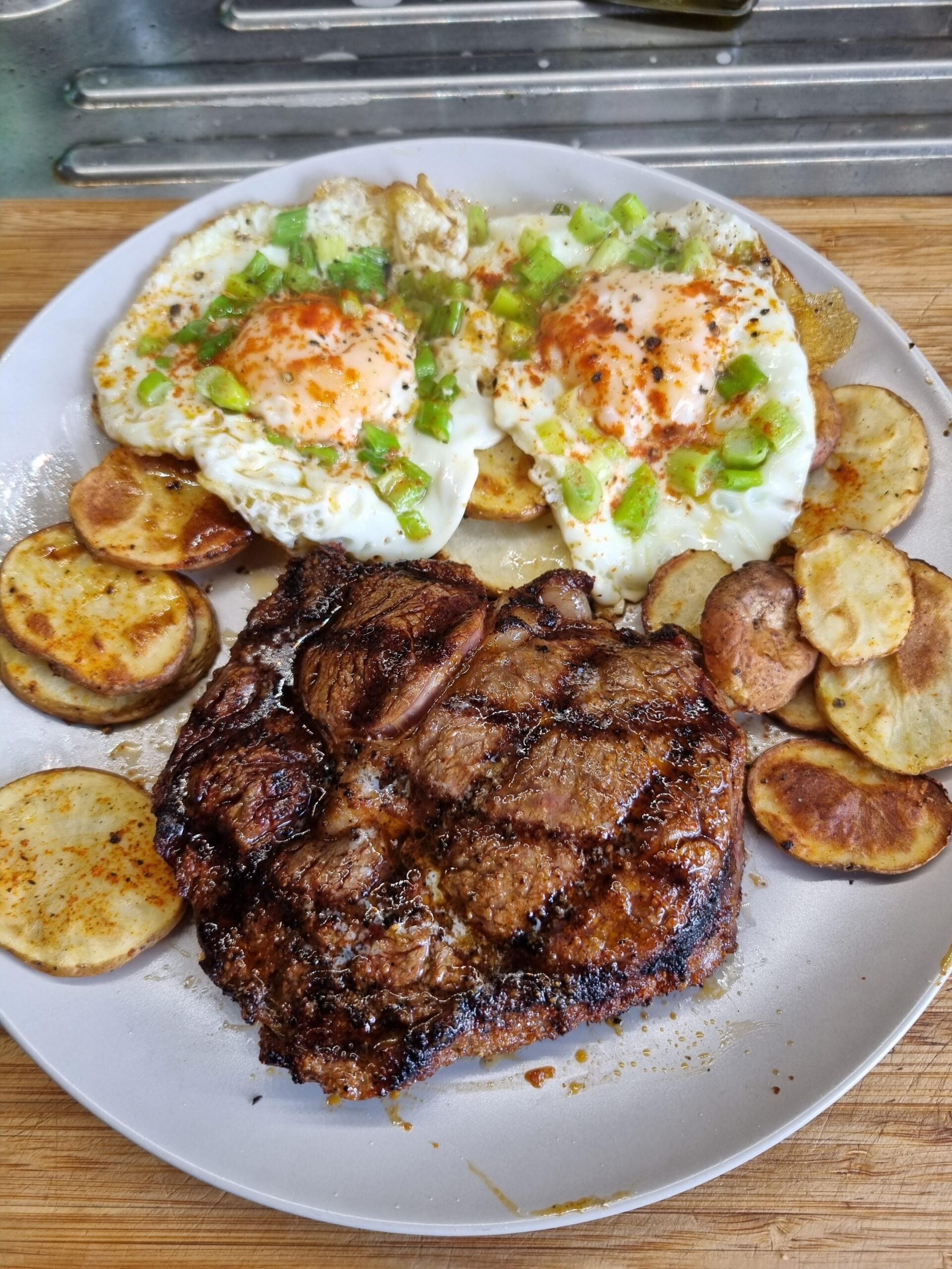 Grilled steak with golden potato slices and two sunny-side-up eggs topped with chopped green onions on a white plate.