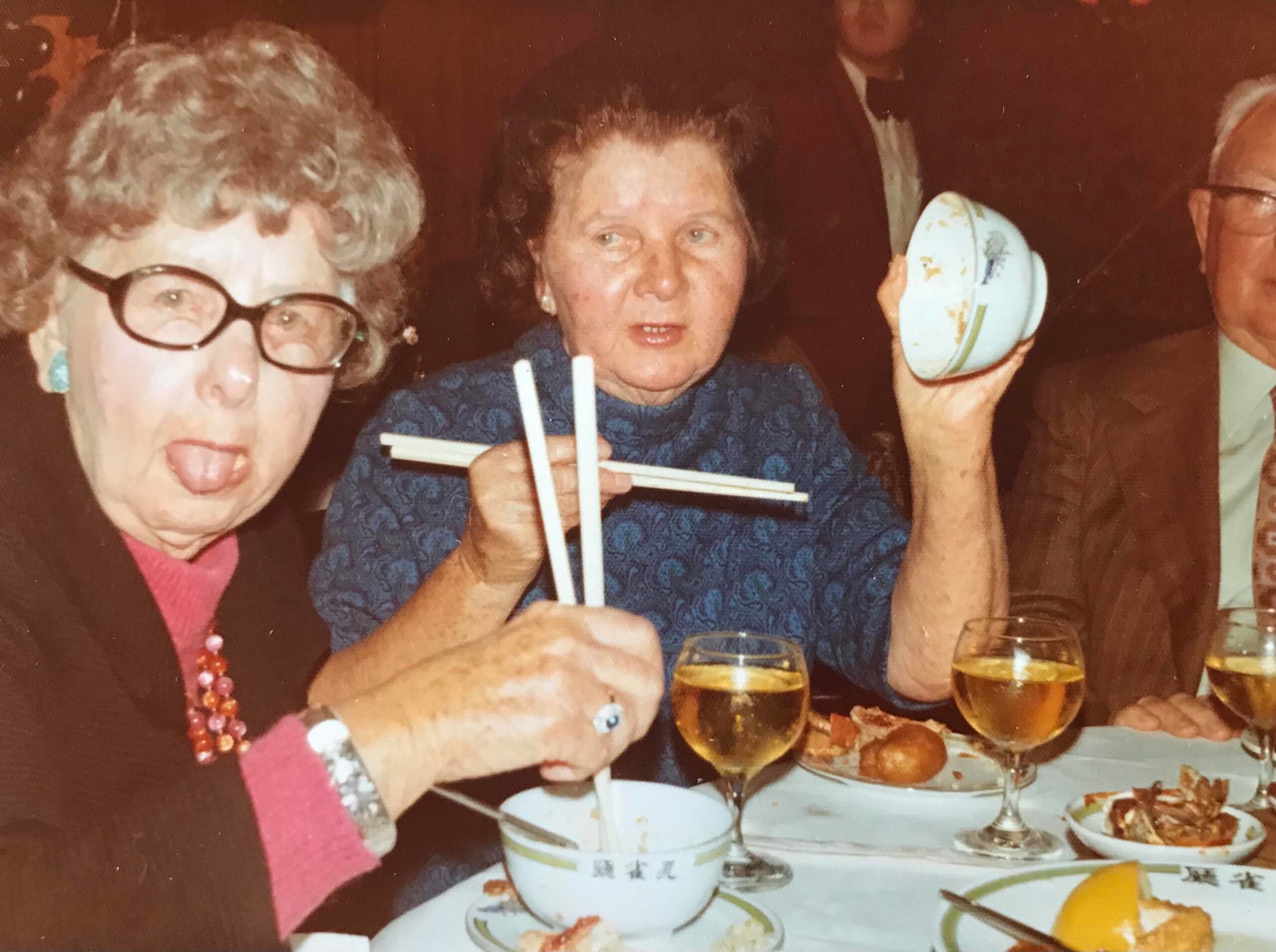Three older adults sit at a table with food and drinks, enjoying nostalgic dinners together; one woman in glasses sticks out her tongue, another holds chopsticks and a bowl, and a man in a suit sits partially out of frame.