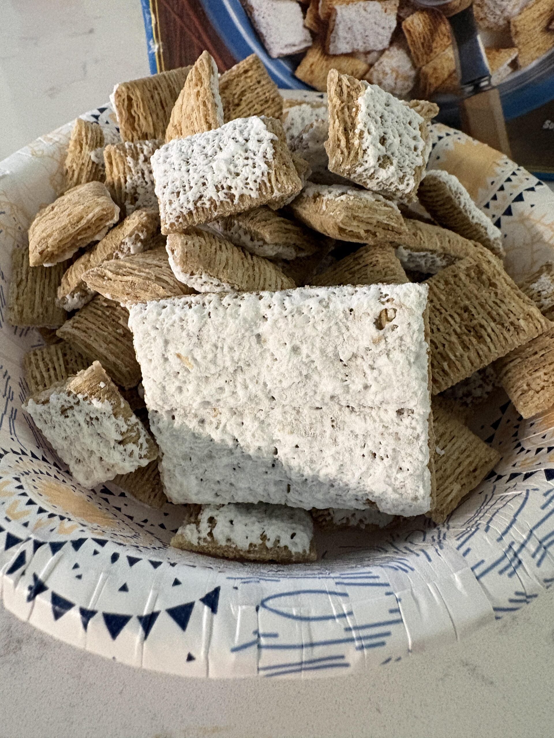 A paper bowl filled with shredded wheat cereal pieces, some coated in a thick layer of white powdered sugar, on a light countertop.