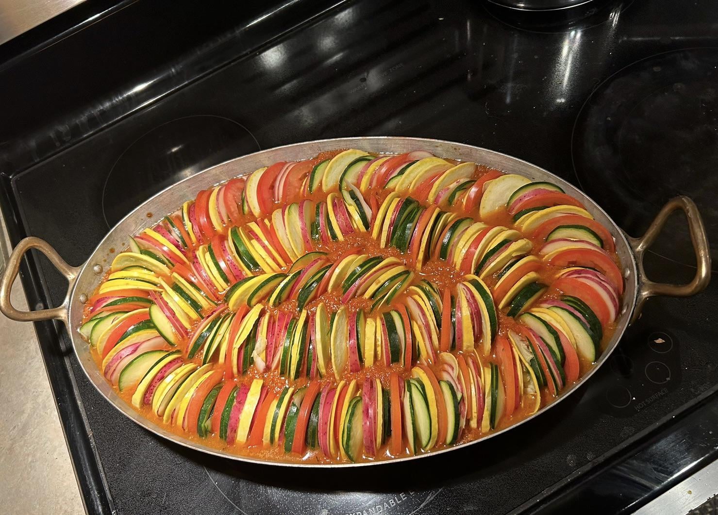 An oval pan of neatly arranged ratatouille with sliced zucchini, yellow squash, eggplant, and tomato in tomato sauce sits on a stovetop, ready to be cooked.