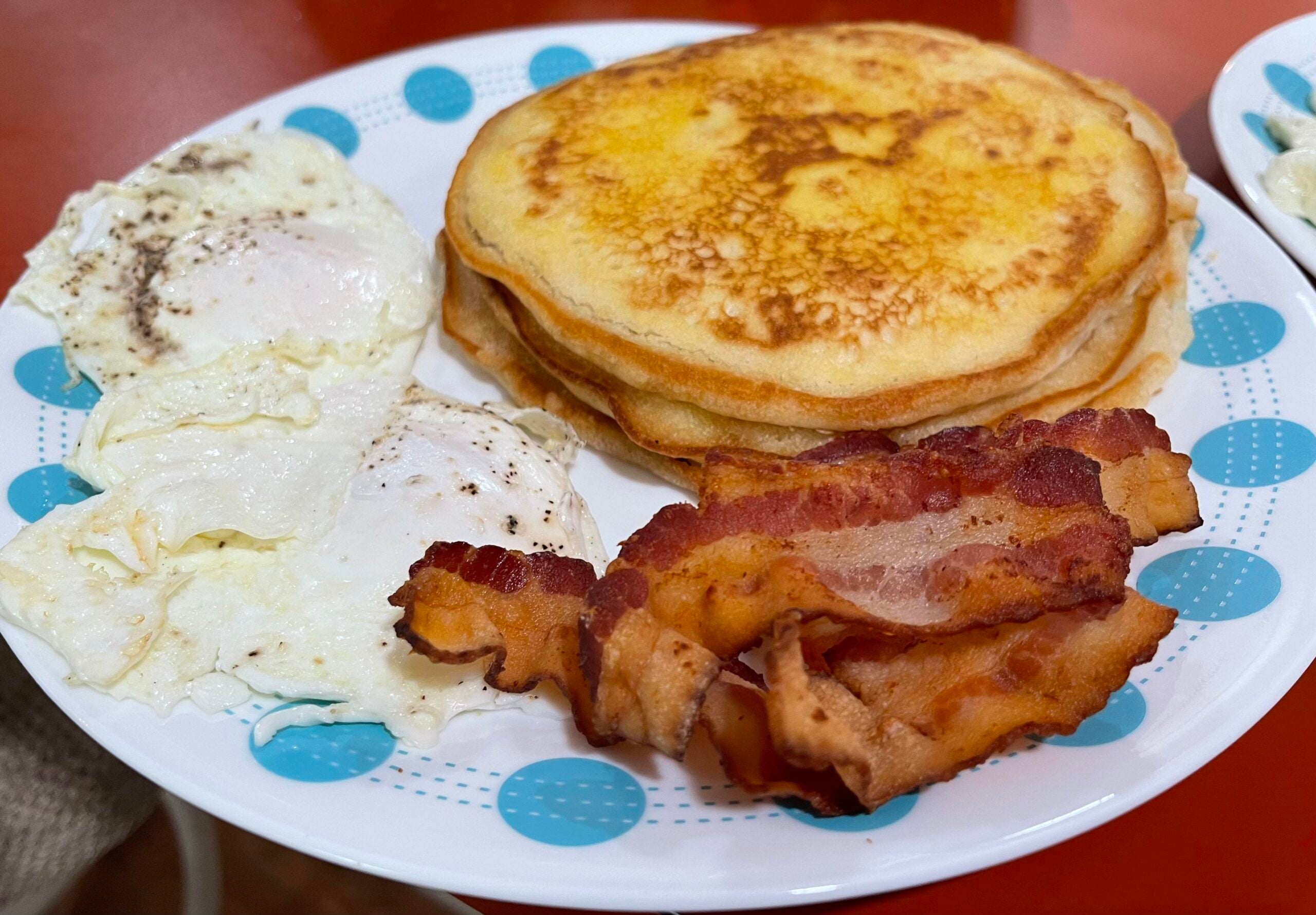 A plate with three golden pancakes, two sunny-side-up eggs, and several crispy bacon strips on a white plate with blue dots.