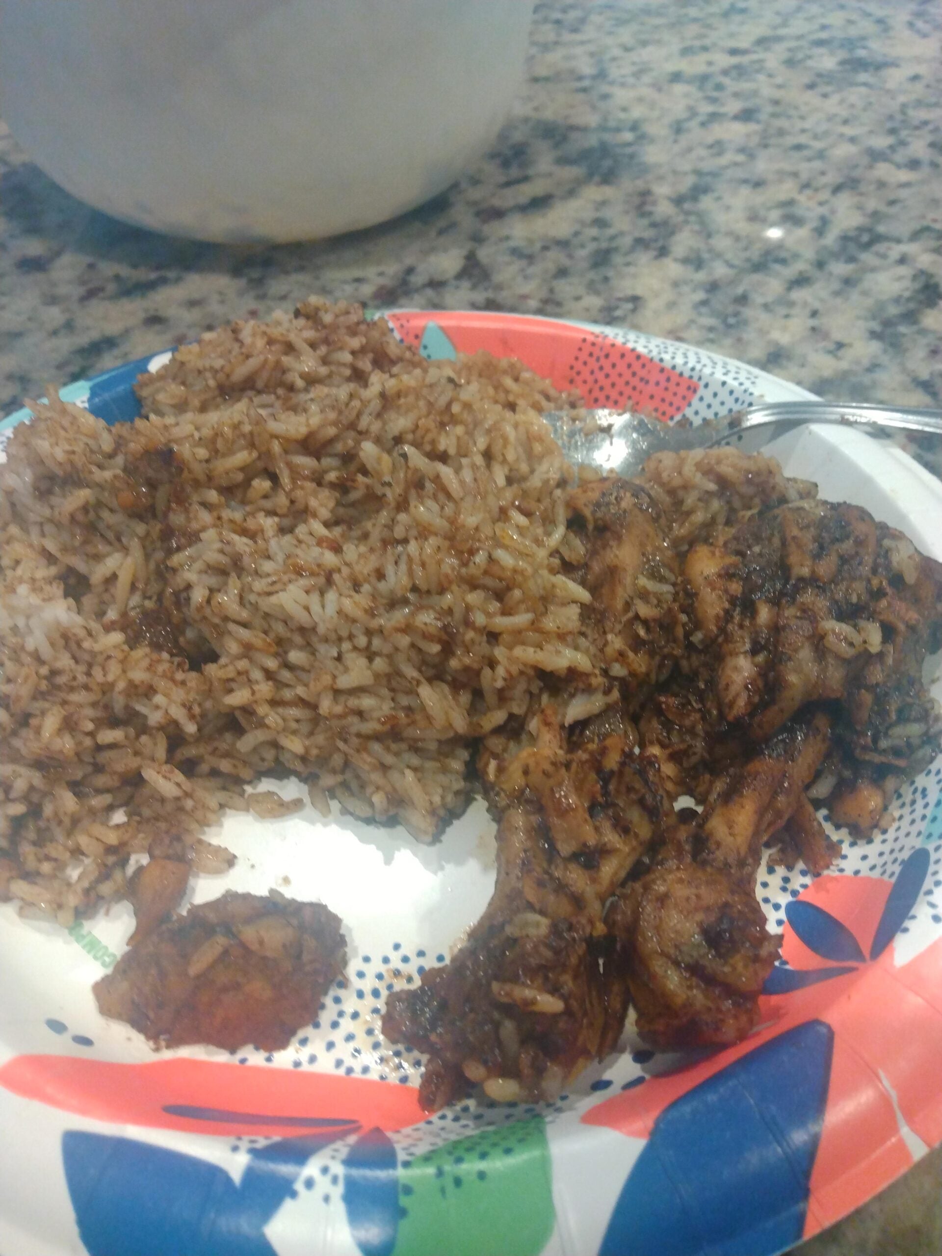 A paper plate with seasoned rice, grilled chicken pieces, and a small serving of fried plantain sits on a granite countertop—a far cry from any hilarious cooking disasters, this meal is perfectly prepared and ready to enjoy.