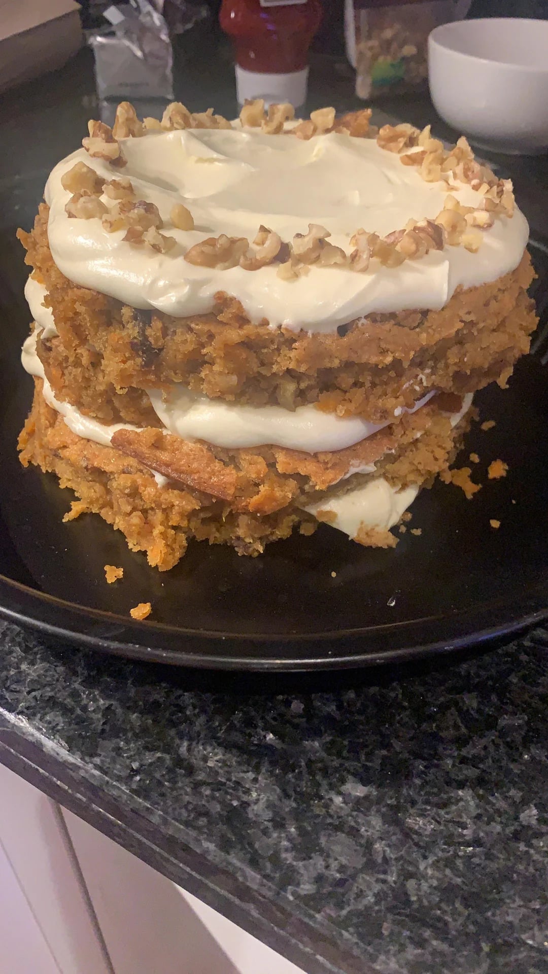 A homemade, uneven layer carrot cake with cream cheese frosting and chopped walnuts on top, placed on a black plate on a kitchen counter.