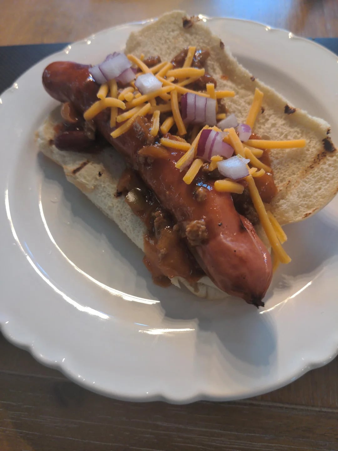 A grilled hot dog in a bun topped with chili, shredded cheddar cheese, and diced red onions sits on a white plate.