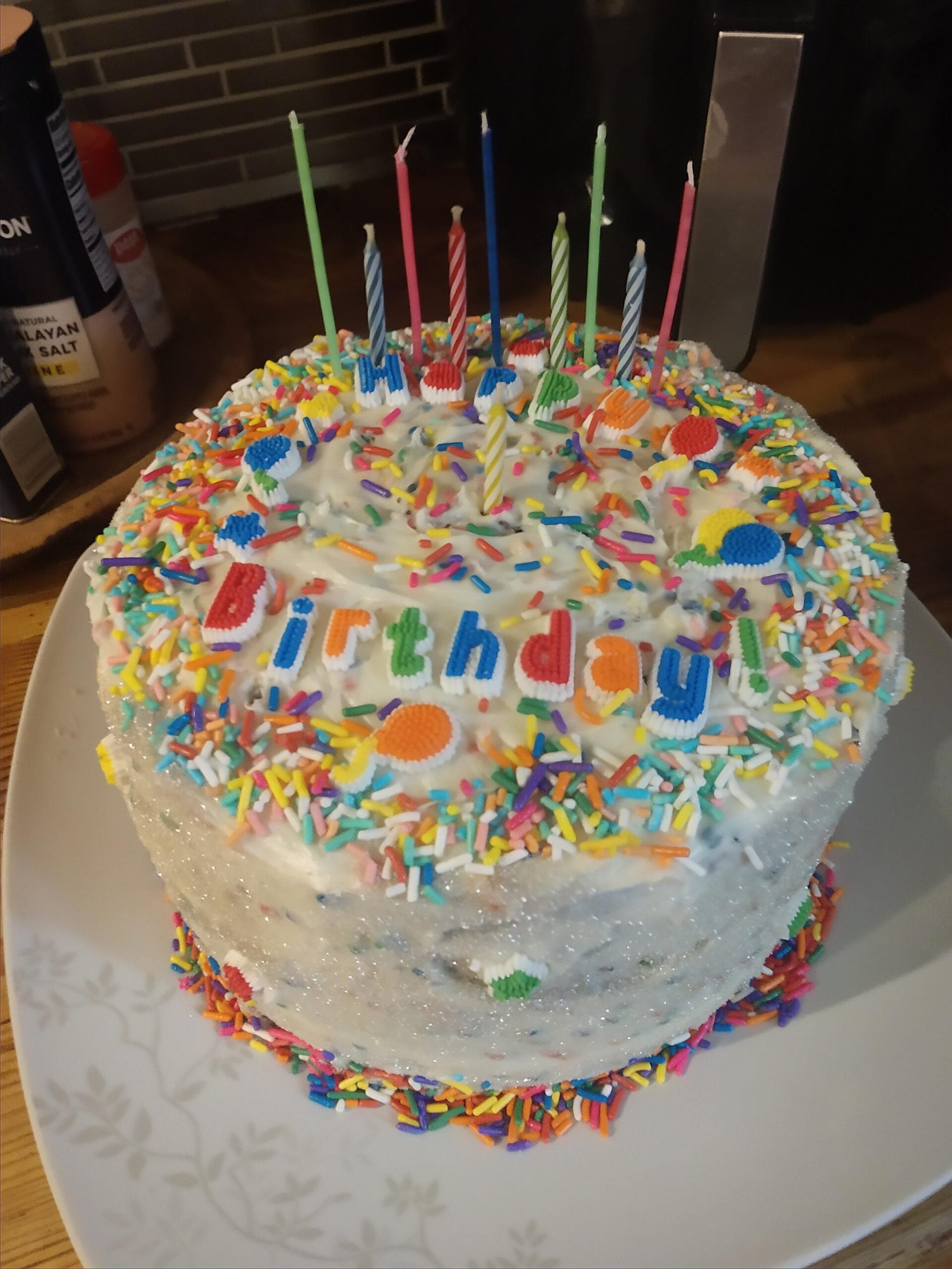 A frosted birthday cake with colorful sprinkles, rainbow candles, and “Happy Birthday!” spelled out in letter decorations. This delightful centerpiece of homemade desserts sits on a white plate on a wooden surface.