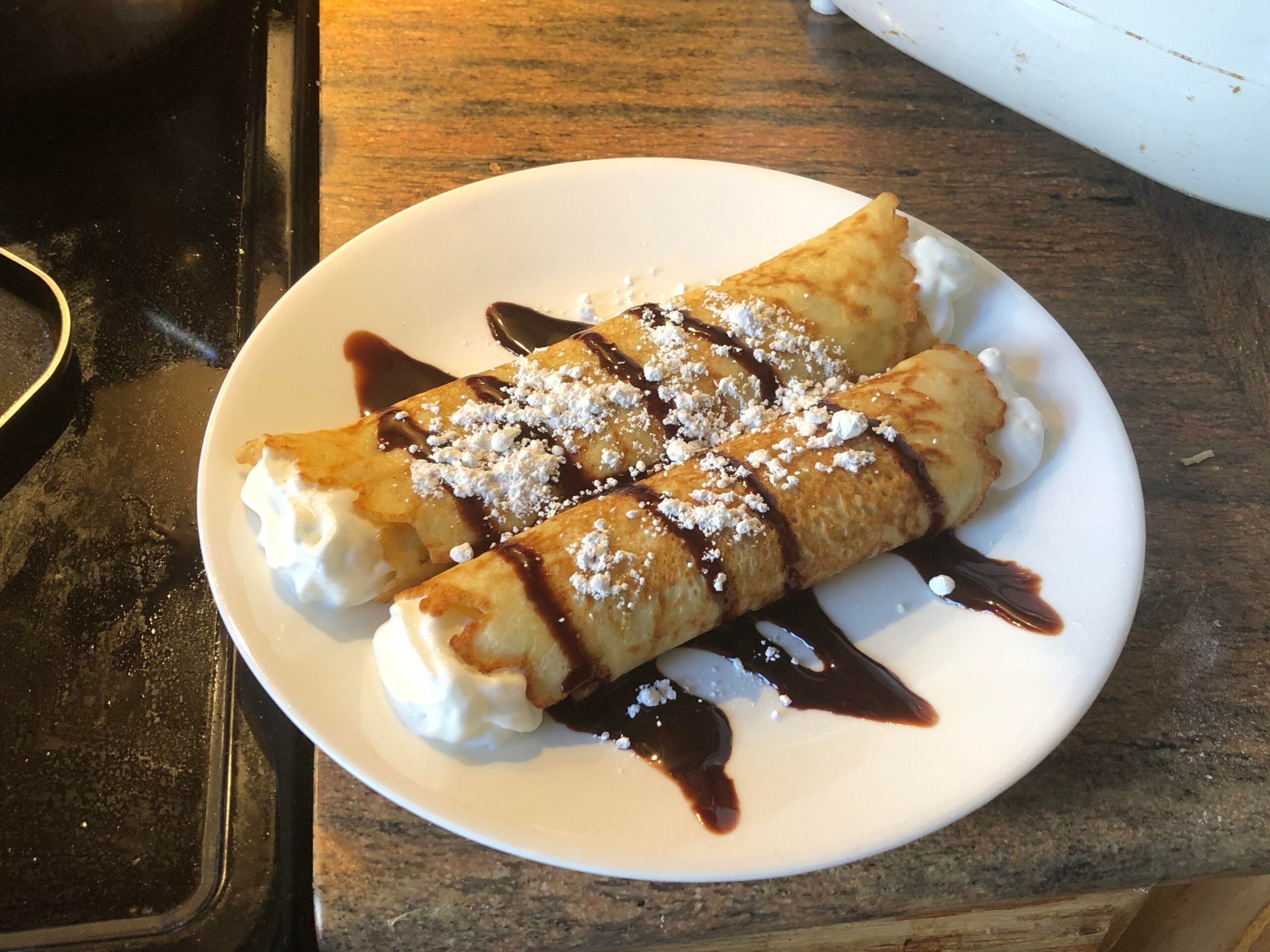 Two rolled crepes on a white plate, filled and topped with whipped cream, drizzled with chocolate syrup, and sprinkled with powdered sugar, placed on a brown countertop—a delectable example of homemade desserts.