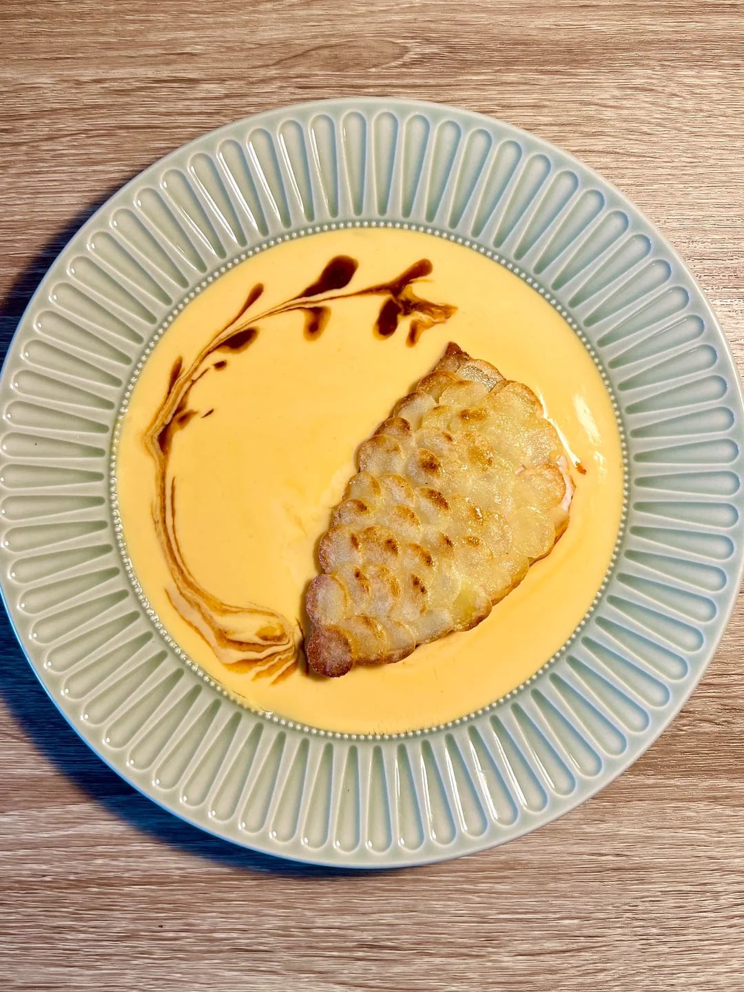 A decorative plate with creamy yellow soup, topped with a fish-shaped piece of food and a brown artistic swirl resembling a branch with leaves. The dish is on a wood surface.