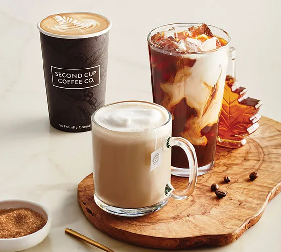 Three coffee drinks on a wooden board, reminiscent of coffee houses: a hot latte in a paper cup with latte art, a glass mug with iced coffee and cream, and a glass mug of hot tea latte; coffee beans and sugar are also visible.