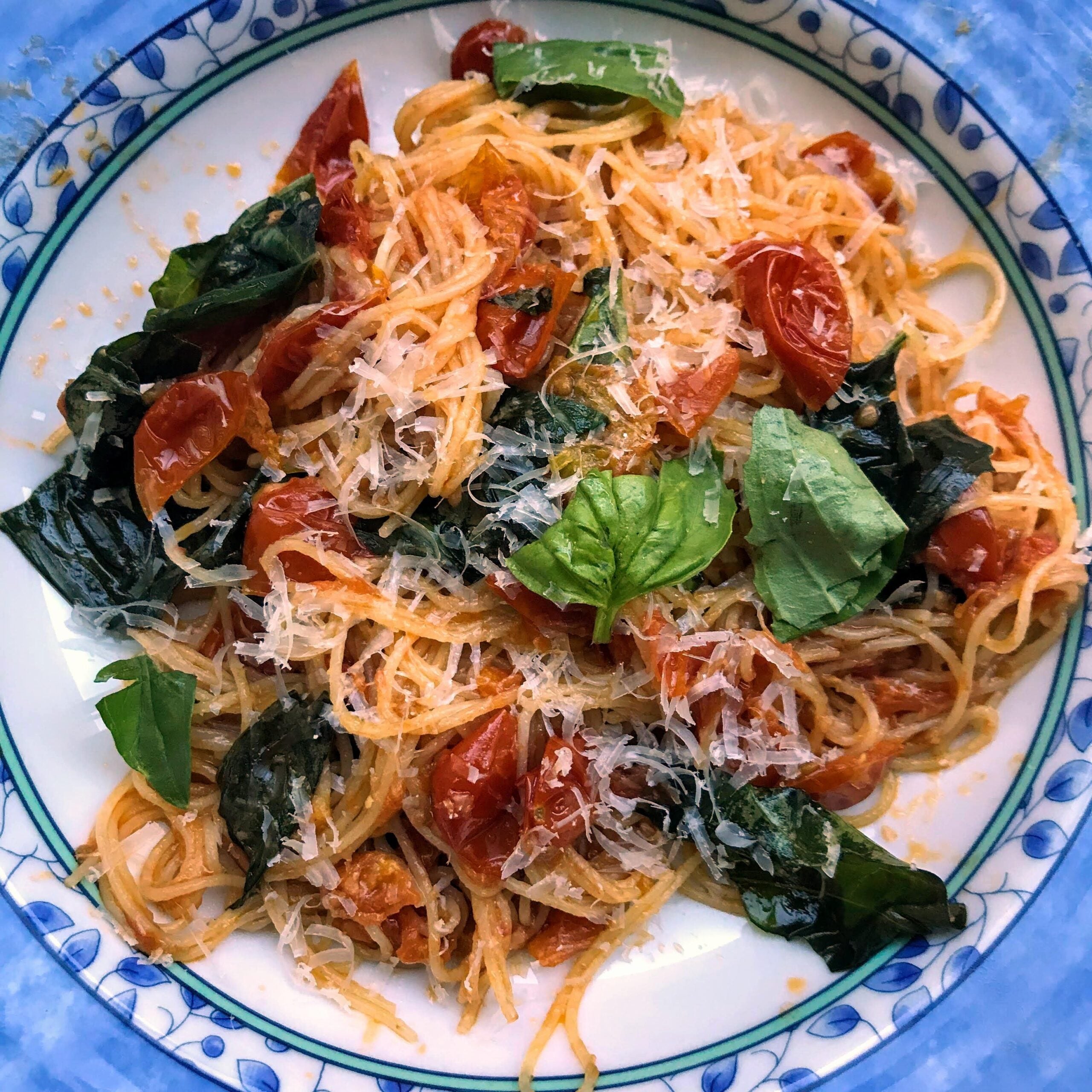 A plate of spaghetti topped with tomato sauce, cherry tomatoes, fresh basil leaves, and grated parmesan cheese, served on a decorative blue and white dish.