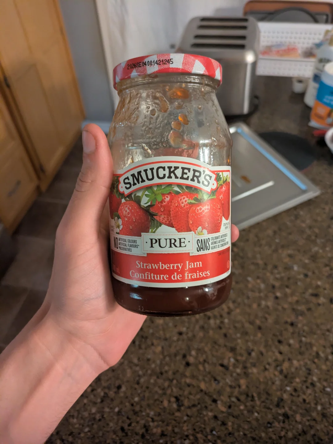 A hand holds a jar of Smucker’s Pure Strawberry Jam in a kitchen. The jar has a red checkered lid and a label with strawberries. A toaster and other kitchen items are on the counter in the background.