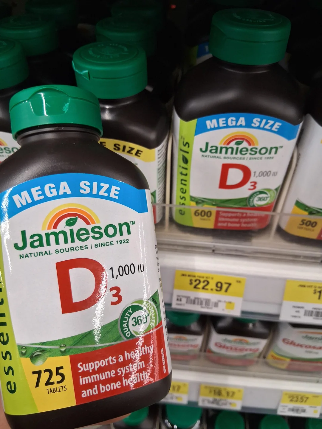 A hand holds a bottle of Jamieson Vitamin D3 (1000 IU, 725 tablets) in front of similar bottles on a store shelf. A price tag below reads $22.97.