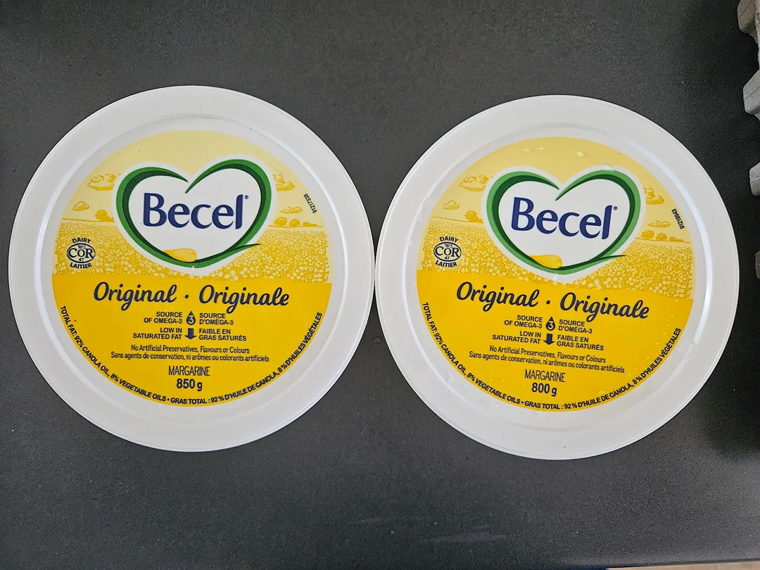 Two round tubs of Becel Original margarine with yellow and white labels sit side by side on a dark surface. One tub is 850g and the other is 800g. Both labels display the Becel logo in a heart shape.