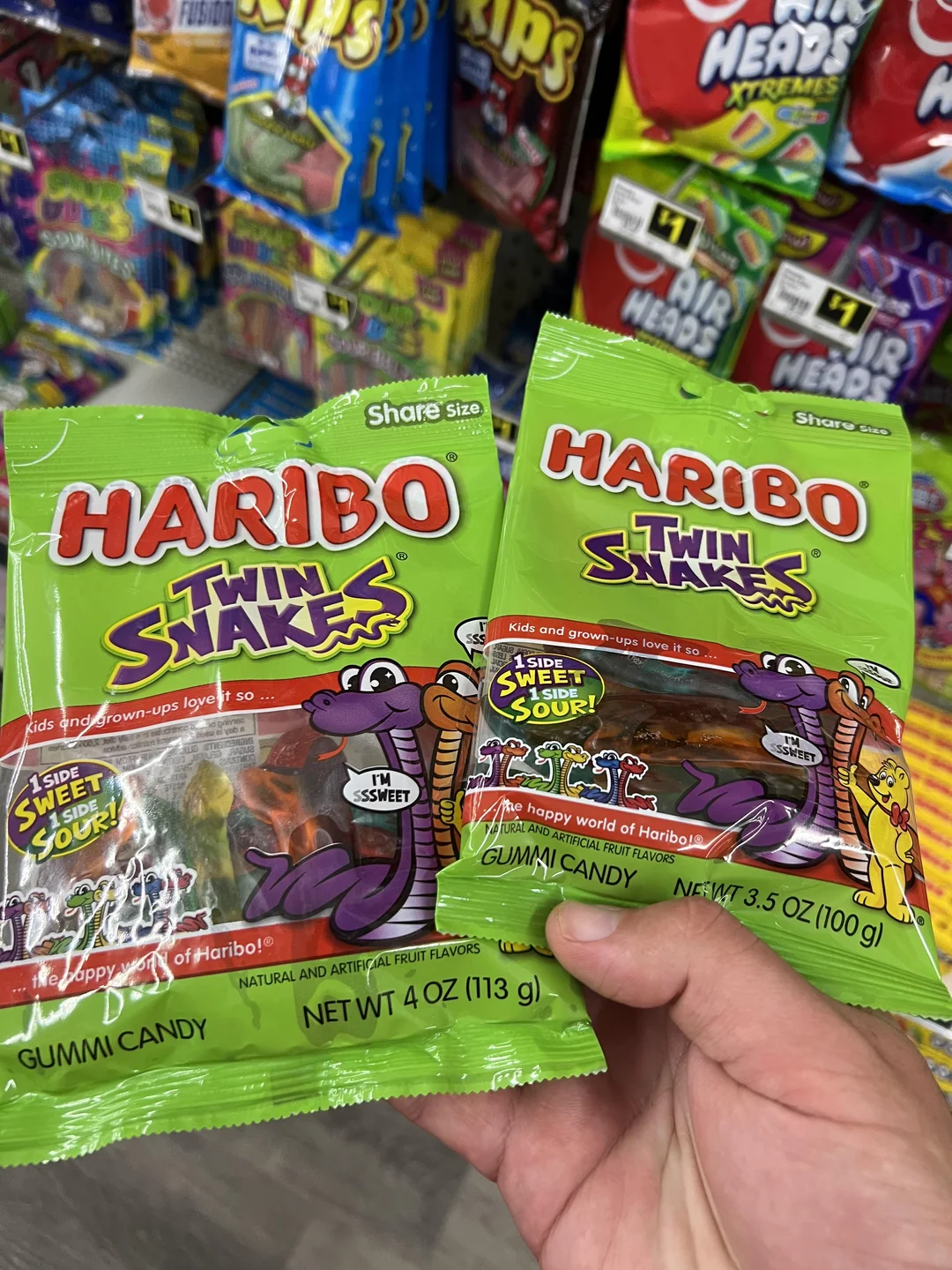 Two hands hold green bags of Haribo Twin Snakes gummy candy in a store aisle, surrounded by other colorful candy on display shelves.