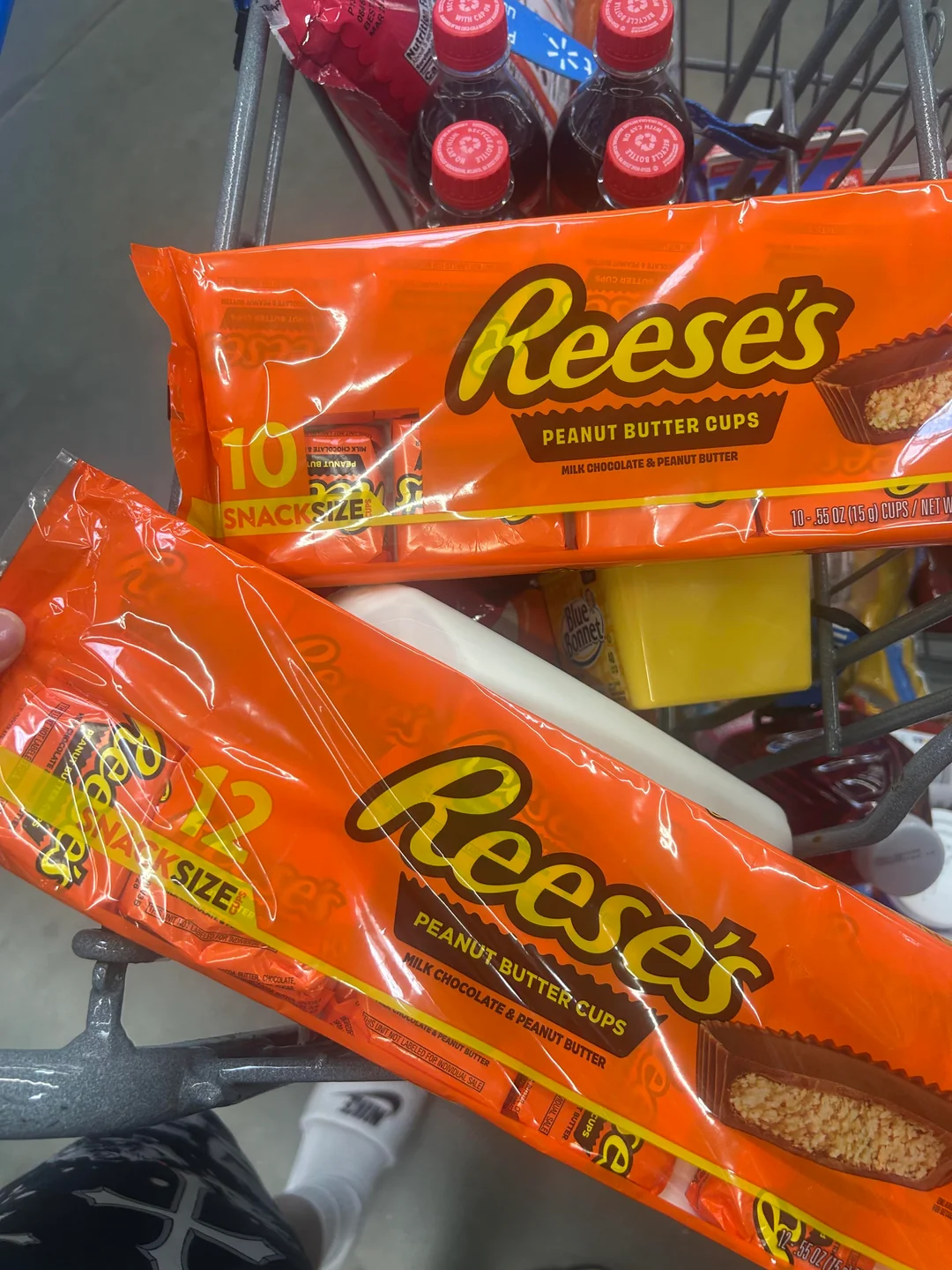 Two large orange packages of Reese's Peanut Butter Cups are being held over a shopping cart filled with drinks and other groceries. The packages are labeled "Snack Size" and "10 Snack Packs.