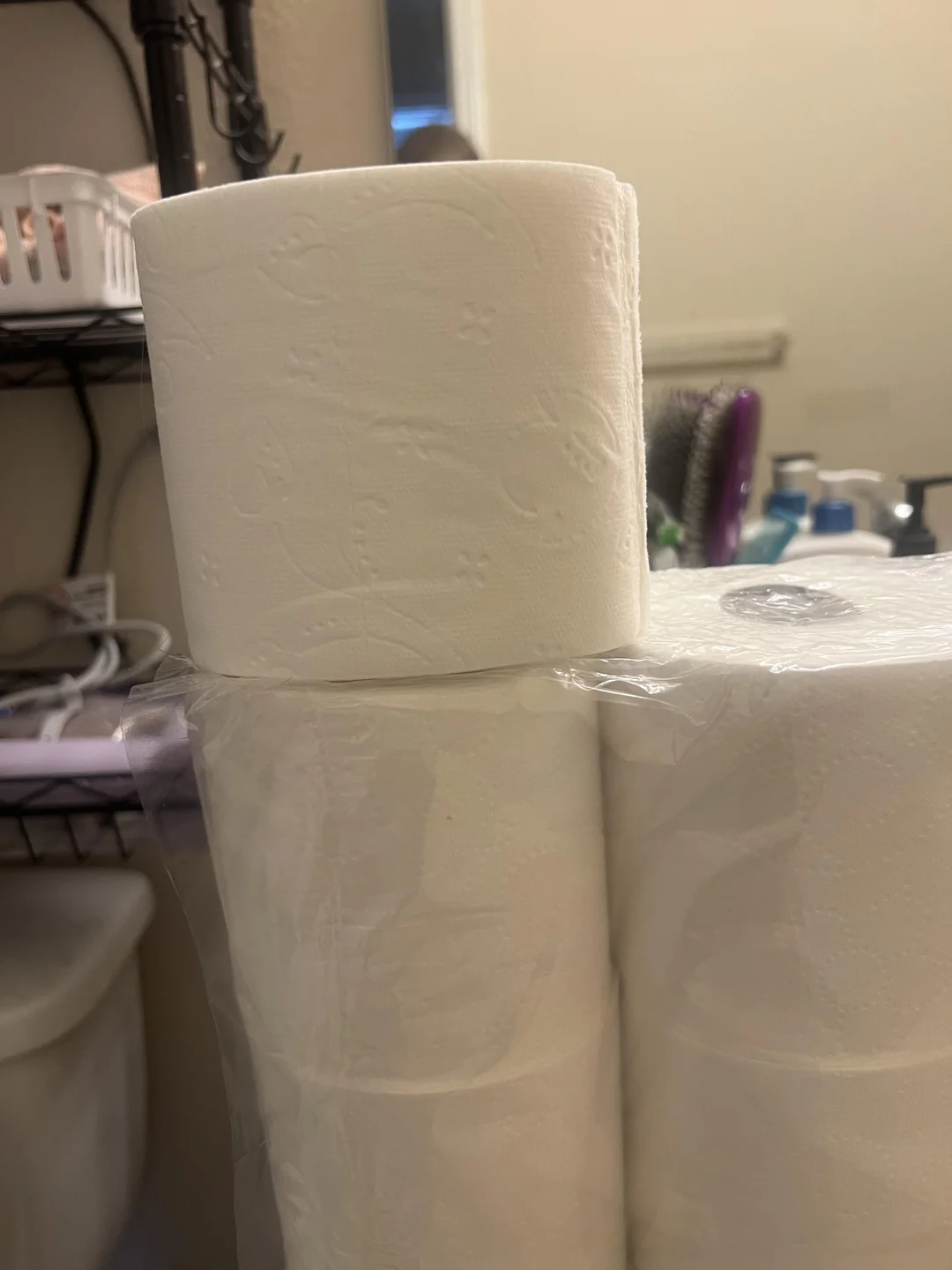 A close-up of a roll of toilet paper resting on packaged rolls in a bathroom. Shelves with assorted items and a mirror are visible in the background.