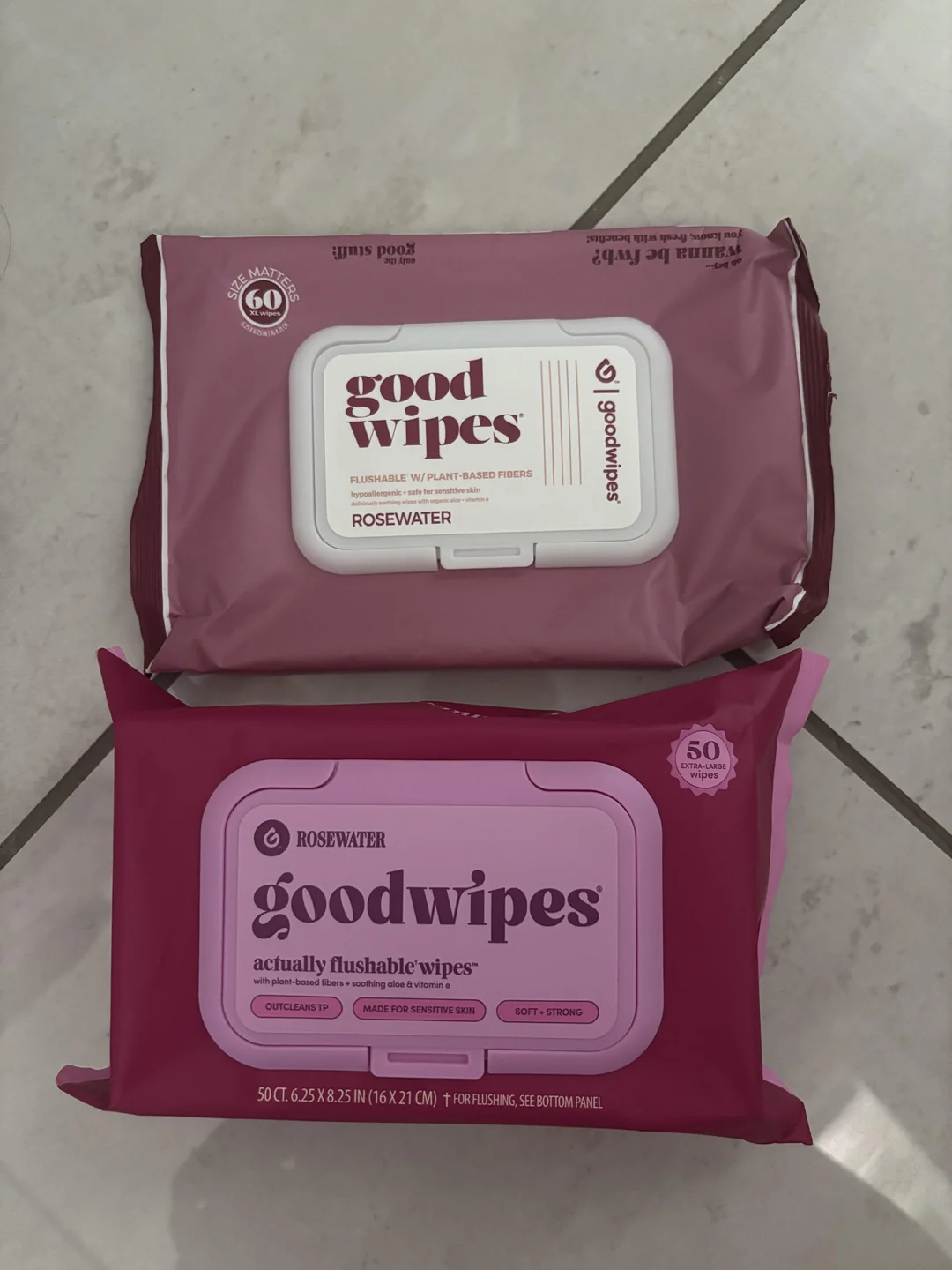 Two packages of Goodwipes Rosewater flushable wipes, one in light purple and one in dark pink, are placed on a tiled floor. Both packages display product details and branding.