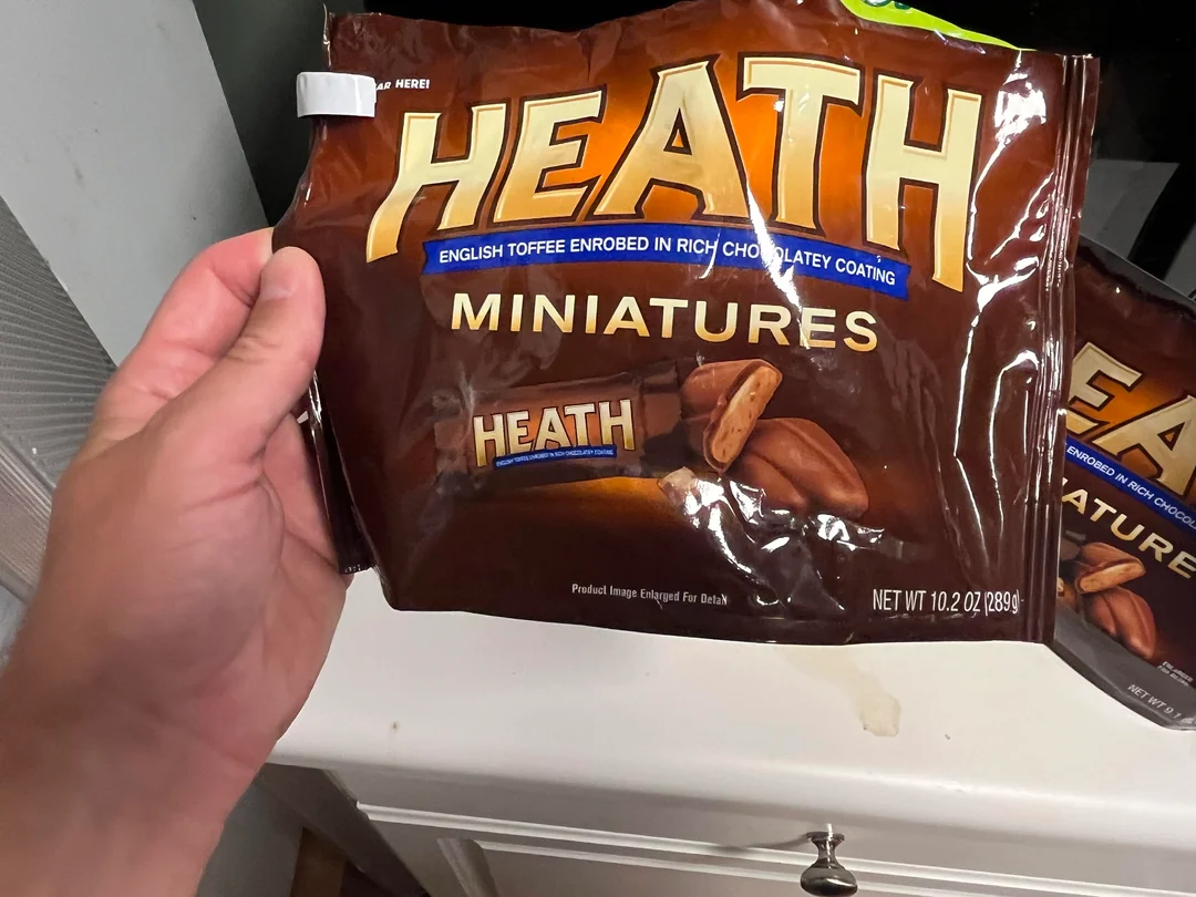 Photos of shrinkflation. A hand holds a bag of Heath Miniatures, English toffee candies coated in chocolate, above a white counter. Another similar bag is partially visible in the background.