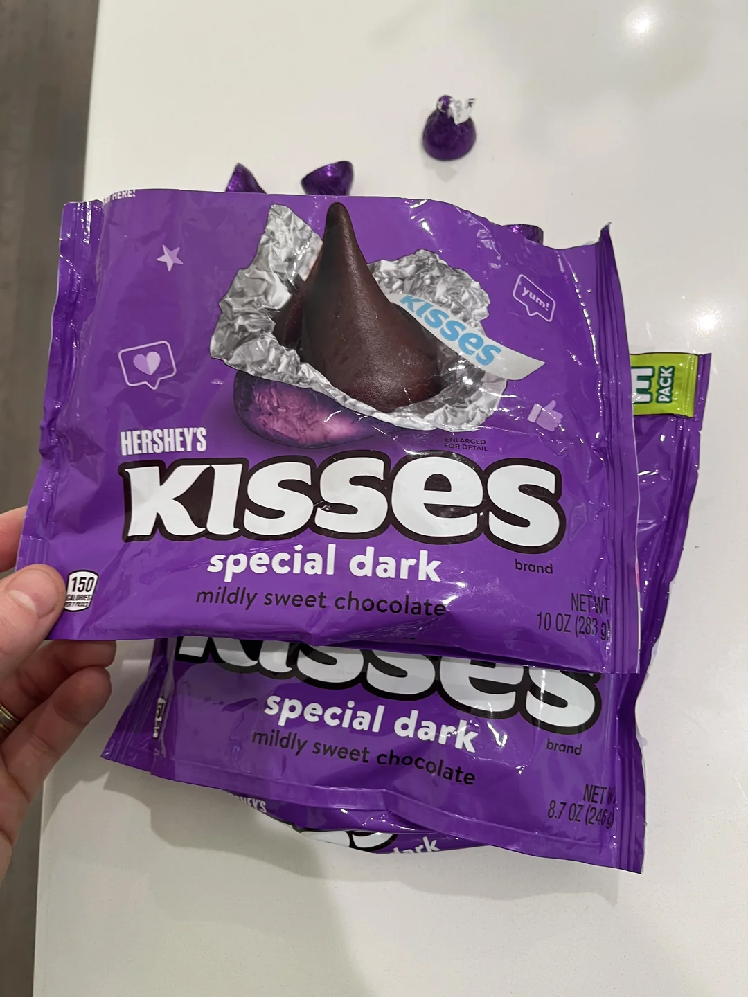 A hand holds a bag of Hershey's Kisses Special Dark mildly sweet chocolate. Two other Kisses and a second bag are on a white surface in the background.