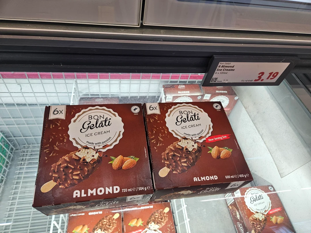 Two boxes of Bon Gelati Almond ice cream bars in a supermarket freezer. A price tag above shows "6 Almond Ice Creams" for 3.19. The boxes feature a picture of a chocolate-coated ice cream bar with almonds and a "New Serving Size" label.