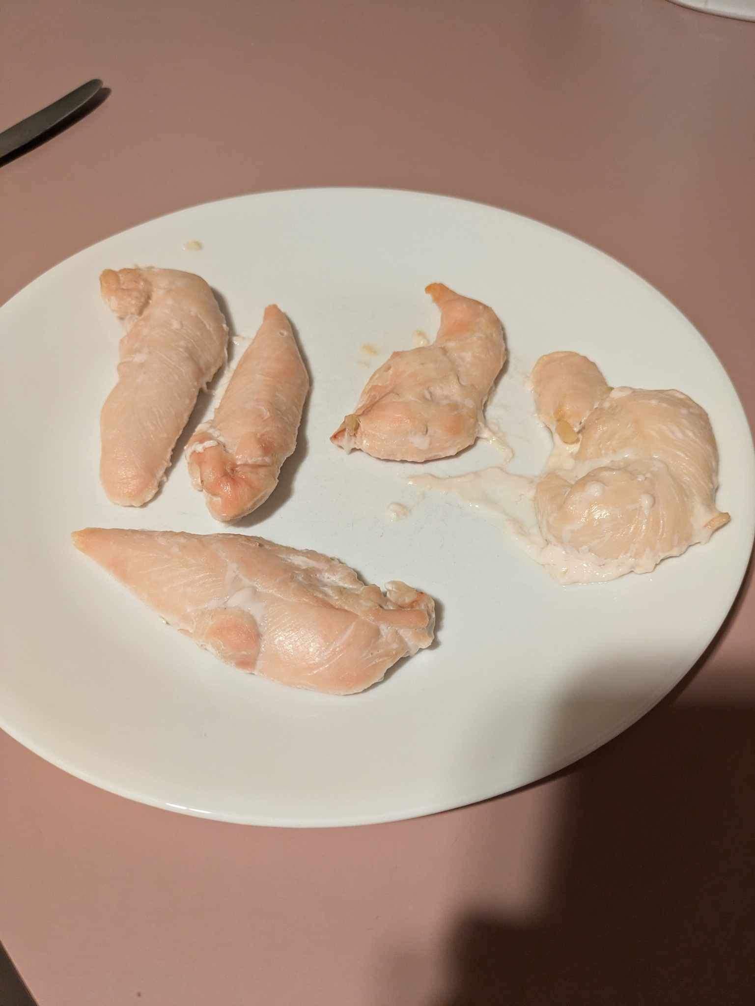 Five pieces of plain cooked chicken breast on a white plate, sitting on a light pink countertop.