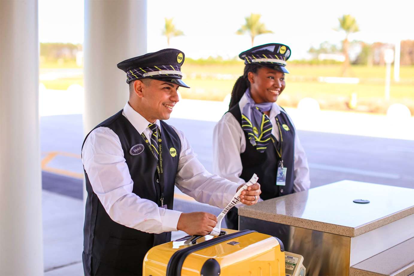 Two uniformed airport staff, a man and a woman, smile as they assist a traveler at an outdoor check-in counter. The man handles a luggage tag near a yellow suitcase, while palm trees are visible in the background.