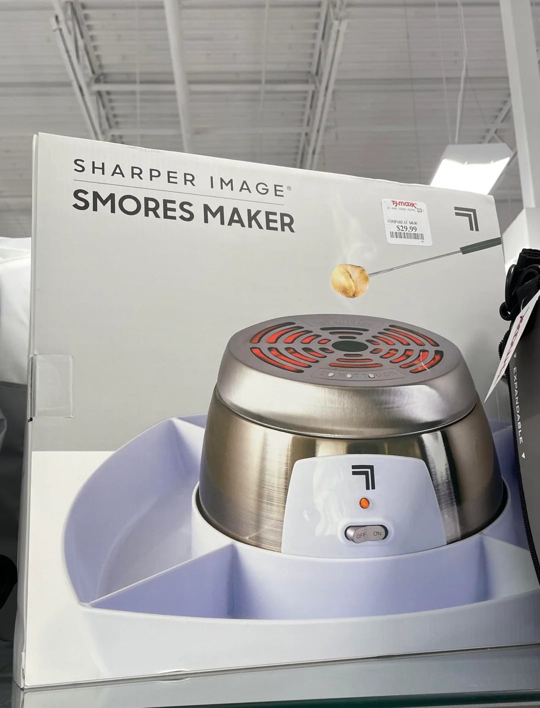 Boxed Sharper Image S'mores Maker displayed on a store shelf; the package shows the appliance with a marshmallow being roasted over its heating element—an innovative addition to your collection of kitchen gadgets.