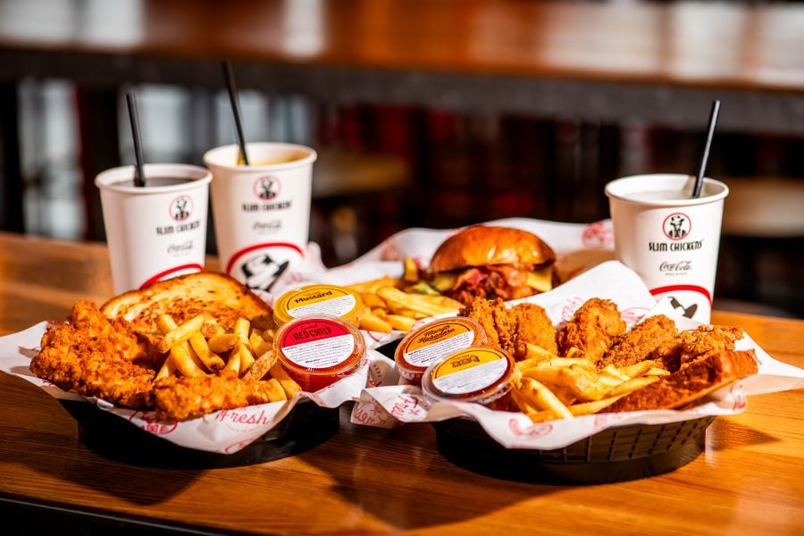 Three baskets of fried chicken, fries, and sauces—reminiscent of popular fried chicken chains—plus a chicken sandwich and three soft drink cups sit on a wooden table in a restaurant.