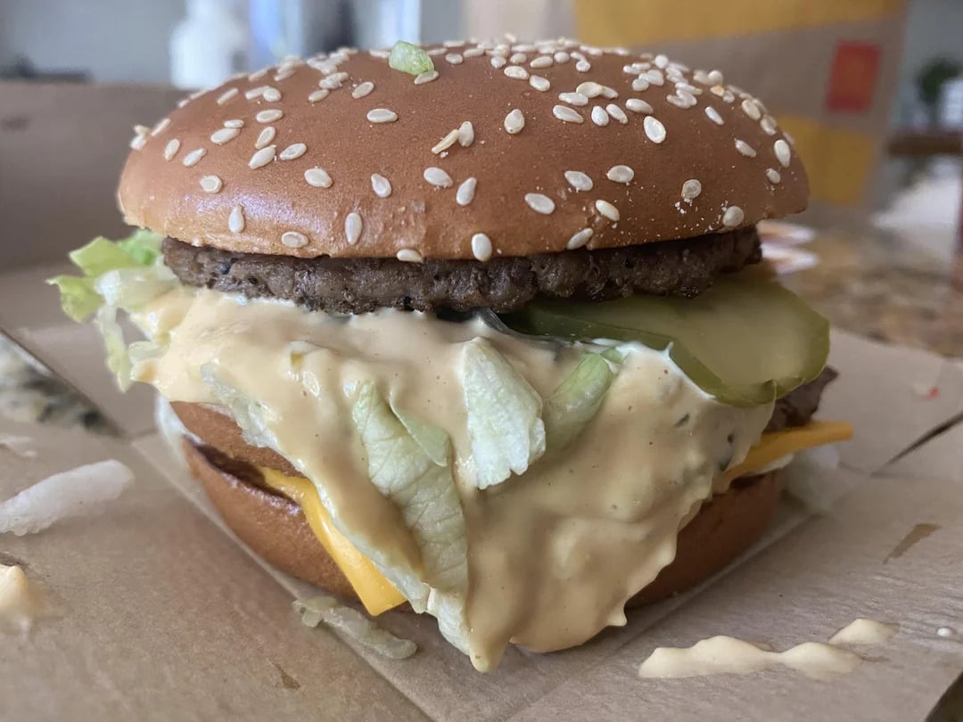 A close-up of a messy cheeseburger with lettuce, pickles, cheese, and sauce overflowing from a sesame seed bun, sitting in a cardboard container—just like you’d find among classic fast-food grocery store products.