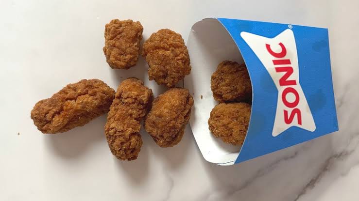 Seven pieces of breaded, fried chicken tenders are scattered on a white surface next to a blue SONIC branded paper container.
