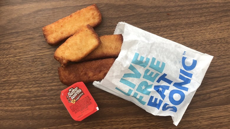 Four fried mozzarella sticks in a Sonic-branded paper bag, with a container of marinara dipping sauce, on a wooden surface.