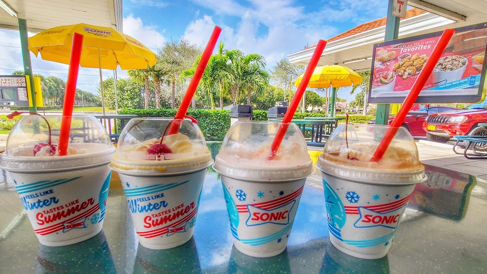 Four Sonic milkshakes with red straws are on an outdoor table under a sunny sky, with yellow umbrellas, palm trees, and a menu board in the background.