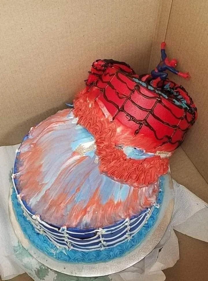 A two-tier cake with red, blue, and white icing, topped with a Spider-Man figure and web design, sits in a cardboard box. The cake is slightly slanted and messy, with Spider-Man appearing to hang from the top tier.