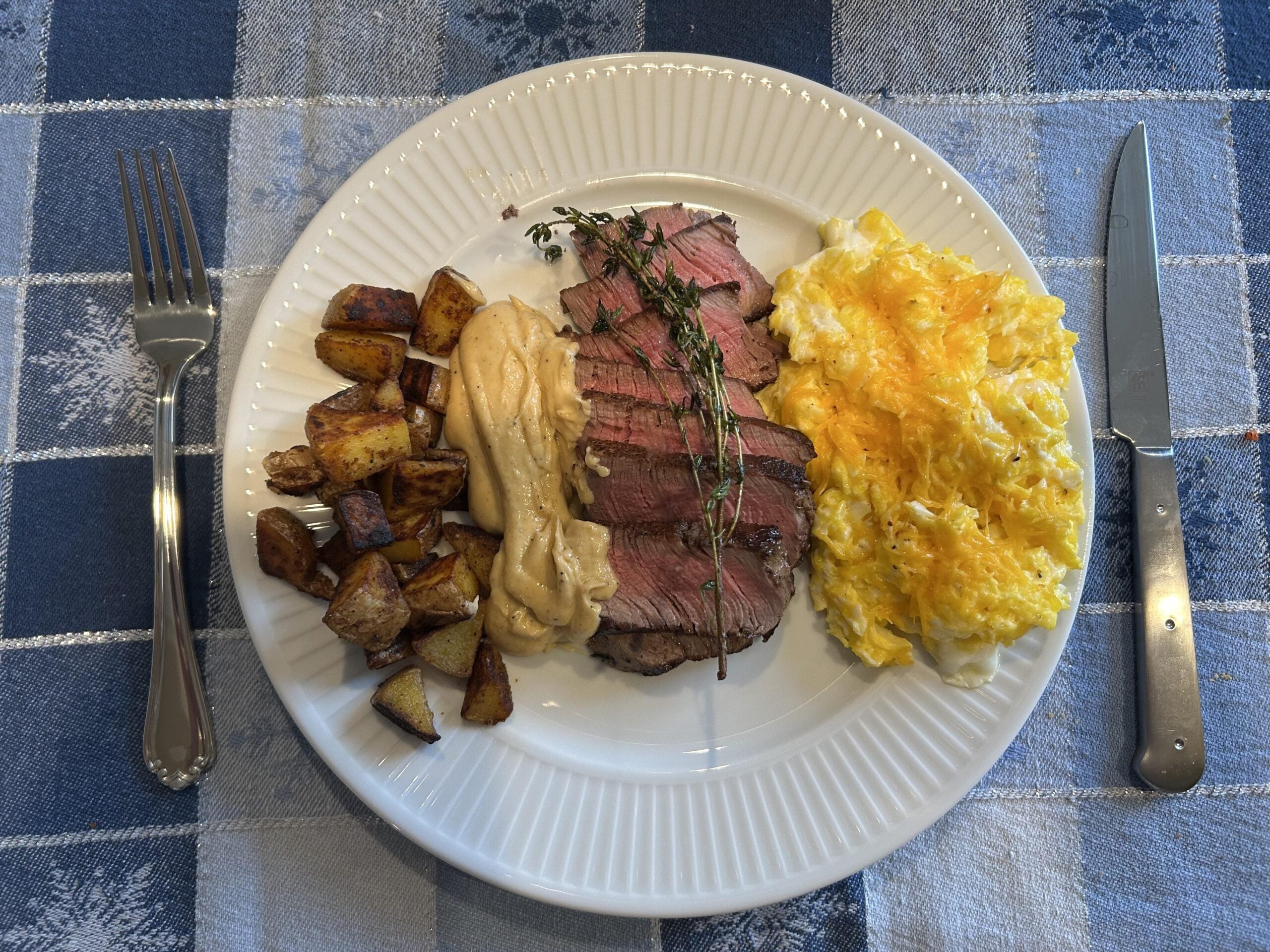 A white plate with sliced steak topped with sauce and herbs, scrambled eggs, and roasted potatoes, set on a blue plaid tablecloth with a fork and knife beside the plate.