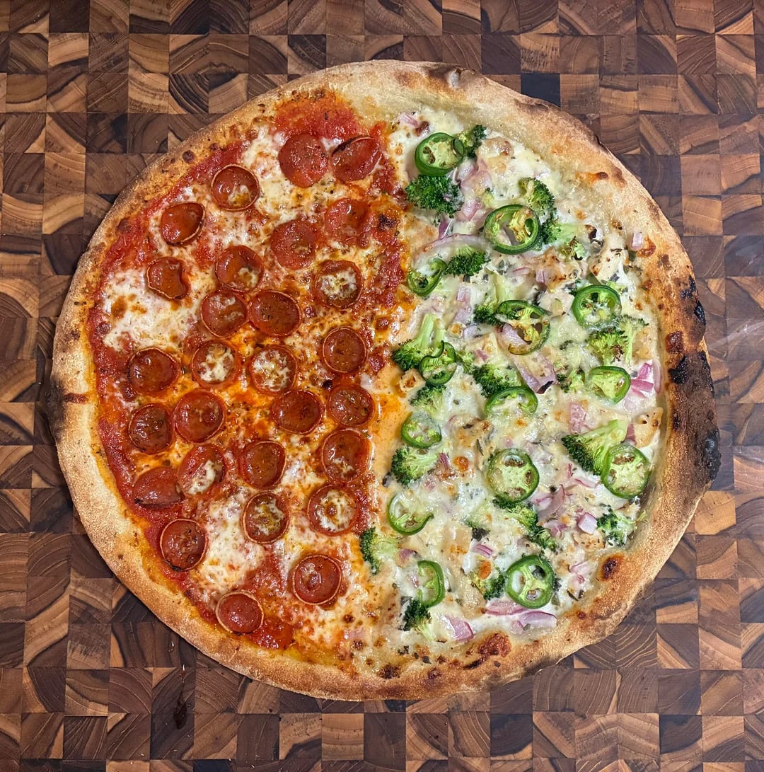 A pizza on a wooden surface, half topped with pepperoni and jalapeños, and the other half with green peppers, onions, jalapeños, and ham. The crust is golden brown and slightly crispy.