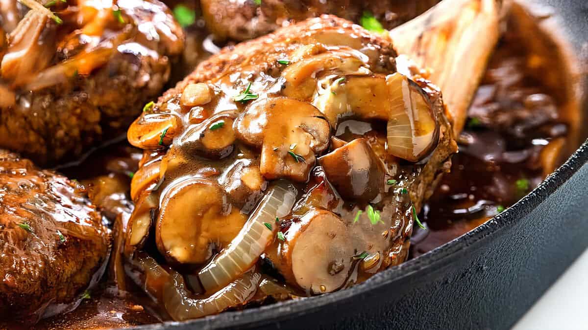 Close-up of a cooked lamb shank topped with sliced mushrooms, onions, and a rich brown gravy in a cast iron skillet.