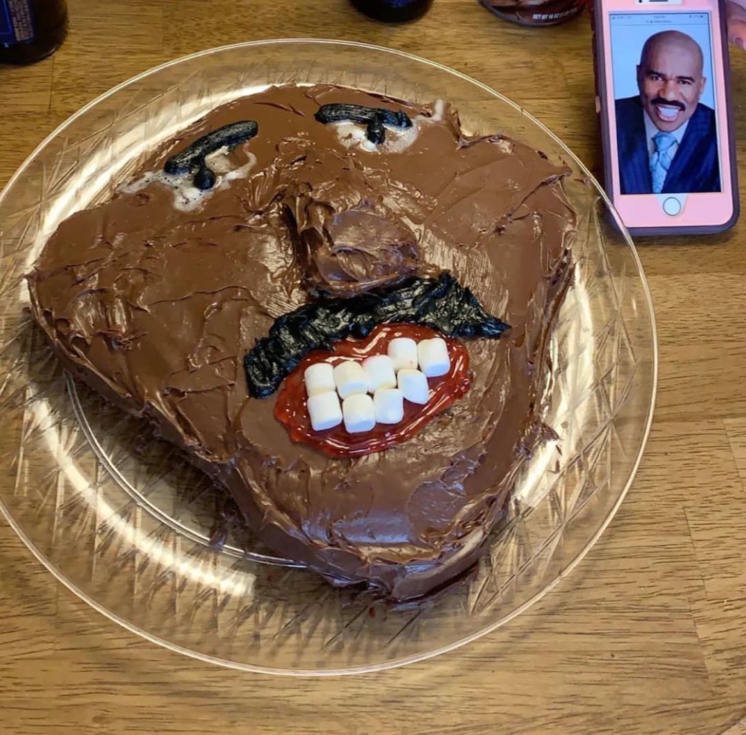 A chocolate-frosted cake shaped like a face with marshmallow teeth and black icing details sits on a plate next to a phone displaying a man's photo for reference.