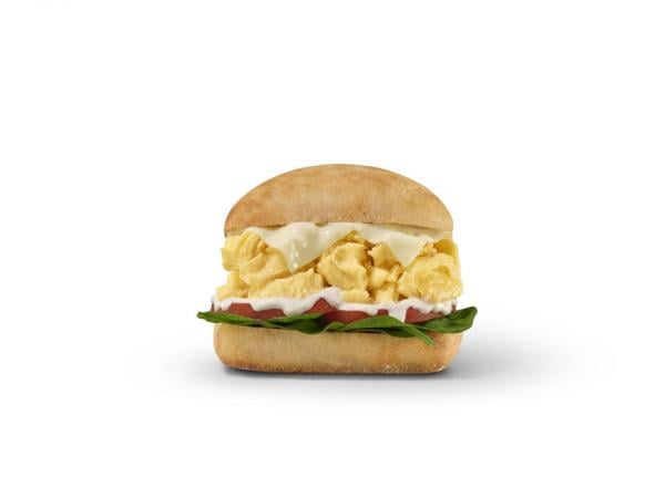A breakfast sandwich with scrambled eggs, cheese, tomato, and spinach on a ciabatta bun, set against a plain white background.