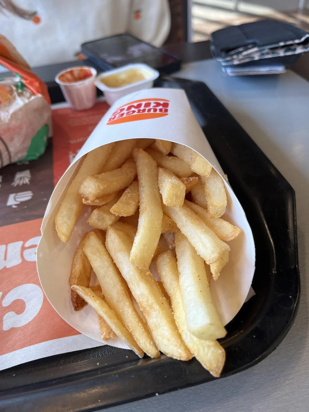 A serving of French fries in a white paper container sits on a black tray with a Burger King logo visible. Nearby are a wrapped sandwich, dipping sauces, a smartphone, and a wallet.
