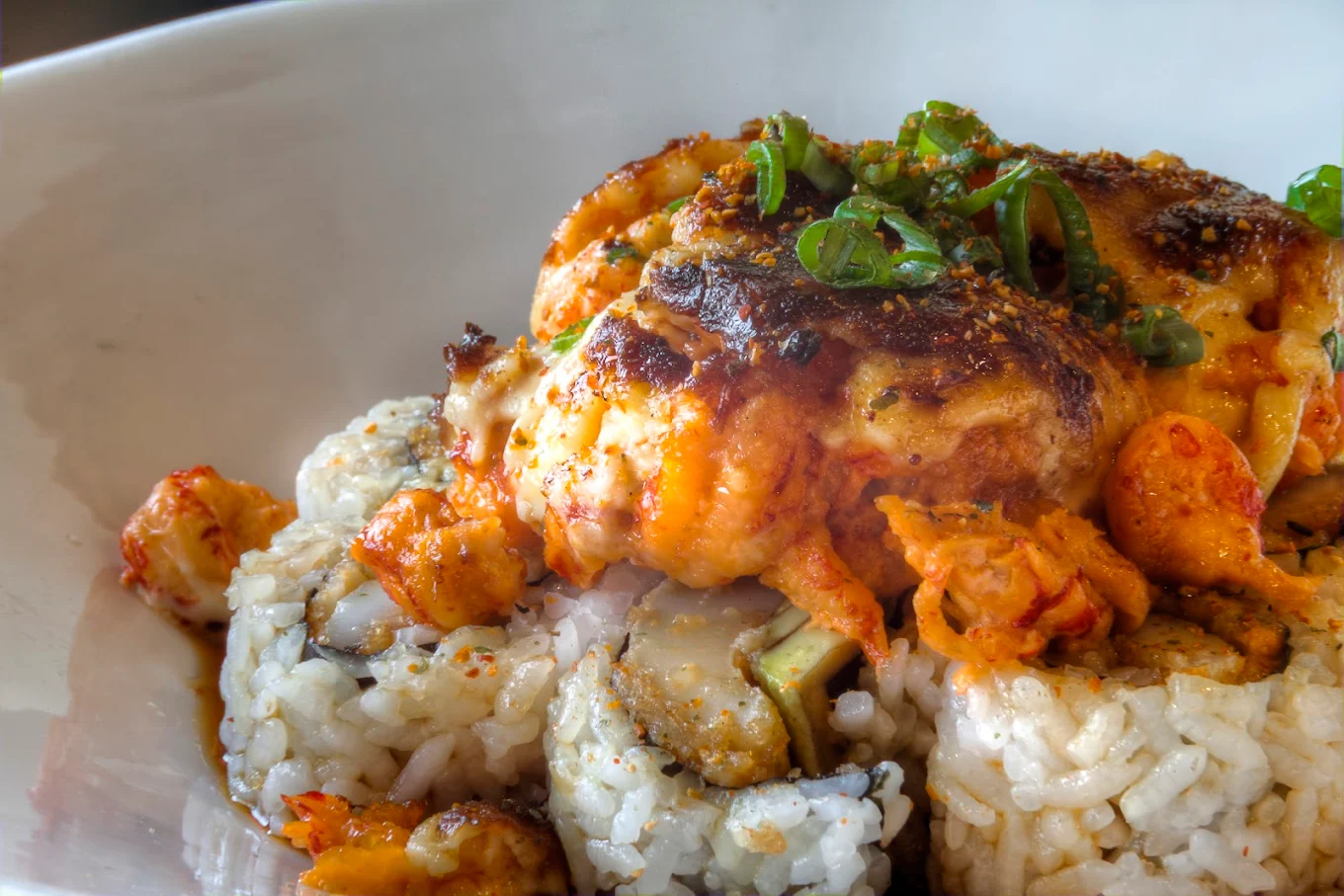 Sushi rolls topped with grilled seafood, creamy sauce, and garnished with sliced green onions, served on a white plate. The filling includes rice and pieces of fish or shellfish.