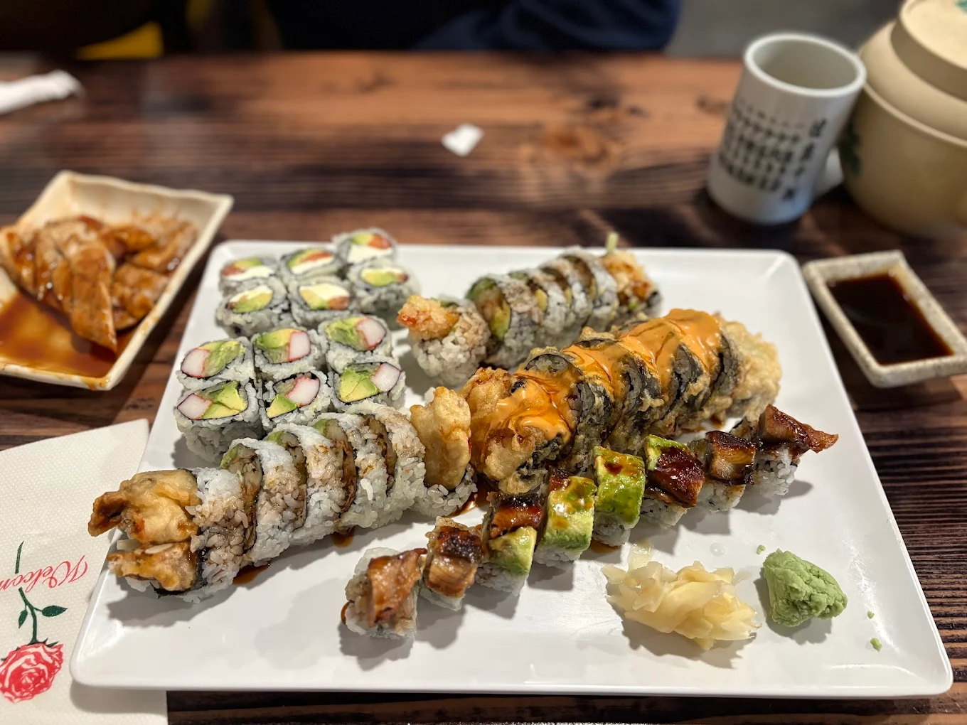A rectangular white plate with assorted sushi rolls, including avocado, crab, and eel, with wasabi and pickled ginger on the side. In the background are gyoza dumplings, a teacup, and a teapot on a wooden table.